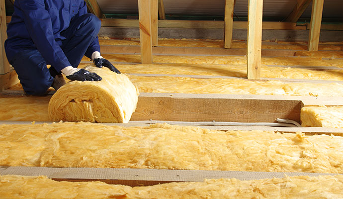 Technician unrolling yellow fiberglass insulation between attic joists